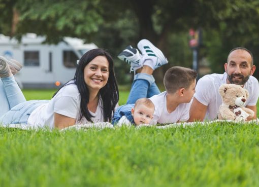 Happy family lying on grass with RV in the background, enjoying a sunny outdoor picnic.
